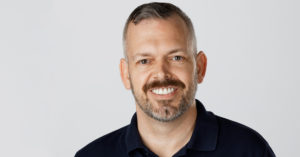 Headshot of Chris Tuttle wearing a black polo shirt and smiling, against a light grey background. Chris is a white guy with salt and pepper hair, a short facial hair, and dark mustache.